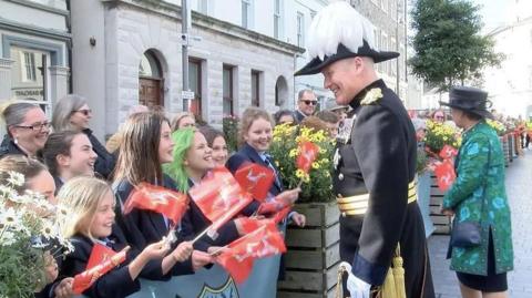 Sir John Lorimer, in his ceremonial uniform, among crowds on the day of his swearing in in Castletown. The uniform in black with gold lapels and there is a row of medals on his chest. His hat is also black and gold with a white plume on top.