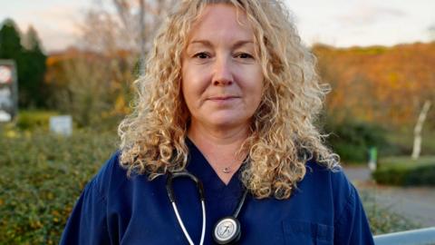 Dr Samantha Jenkins wearing blue scrubs and with curly blonde hair looks at the camera