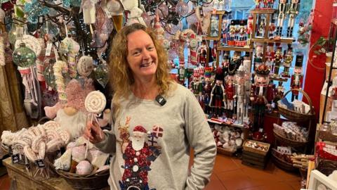 A lady with blonde hair with a grey jumper with a red, white and black sparkly Santa Claus and a brown, red and creme, sparkly reindeer holds a sparkly lollipop in front of a colourful shop full of Christmas decorations