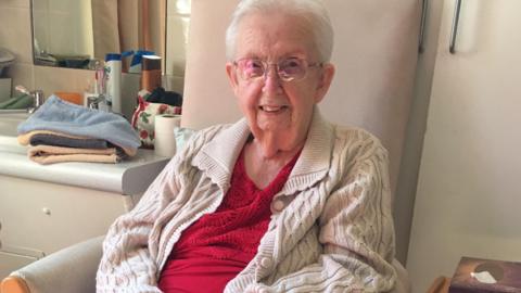 Brenda McNamara is elderly and sits in a chair in a care home. She has silver short hair, clear framed glasses and wears a wool cream-coloured cardigan and a red top. 