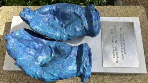 The Nurse's Hands sculpture viewed from above. It is a pair of cupped hands formed out of a blue material. There is a silver-coloured plate below it with the inscription: The Nurse's Hands 2024 Recycled PPE By Kurt Jackson Dedicated to those who care and those we lost