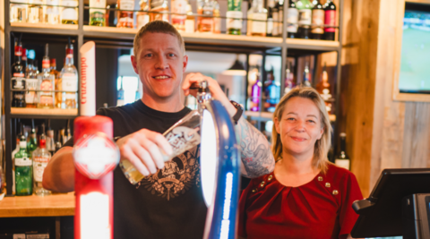 A man with short hair on the left is wearing a black top and standing next to a woman with brown and blonde hair who is wearing red. They are standing behind the bar and bottles are on shelves in the background.