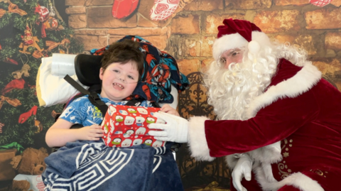 A young child in a mobility wheelchair smiles at the camera whilst being handed a present by a man dressed as Santa. 