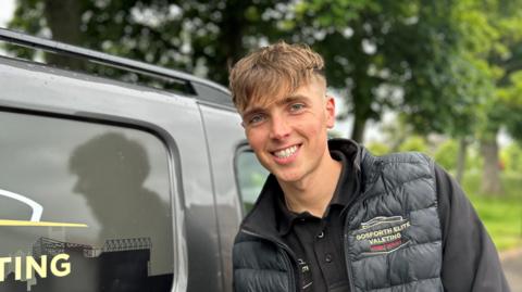 Louis Clougher has brown hair which is longer on top and cropped to the sides and wears a black padded gilet with his company's branding. He is leaning against one of his valet vans and smiling. 