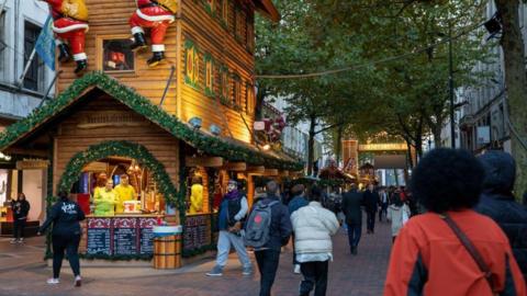 Birmingham Frankfurt Market in the day time. Wooden huts can be seen with the nearest showing two people selling food. Two model Father Christmases are climbing up the side of the hut. Crowds are walking by and there is a large green tree on the right and some shops in the background.