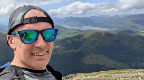 A man wearing green-tined sunglasses, a black cap on backwards and a grey T-shirt is standing on a mountain. In the background is a view of rolling hills and a reservoir.