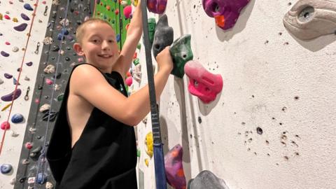 A boy with short blonde hair and wearing a black, sleeveless top, smiles as he holds onto handholds while scaling an indoor climbing wall.