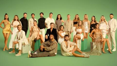 The cast of The Only Way is Essex in front of a green backdrop. Twenty-one of the cast are standing and the rest are sitting either on green seats or on the floor.