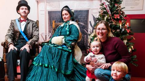 A man and a woman on the left dressed in Victorian costumes sitting on Victorian chairs next to a Christmas tree on the right. In front of a tree is a woman holding a toddler and next to her is another small child