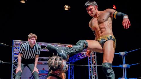 Two wrestlers, one kicking the other in the head, in a wrestling ring while a young referee watches on