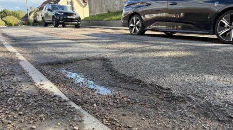 Pothole in a surburban road with two black cars parked by the kerb