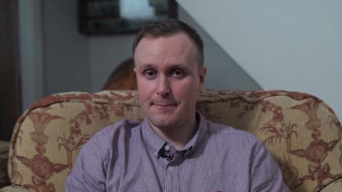 A man with short brown hair is wearing a purple shirt looking into the camera. He is sat in a large yellow/cream arm chair with red designs on it. There is a blue/white wall in the background.