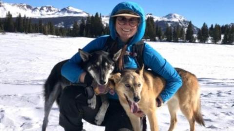 Emilie Anne Parker looking directly at the camera lens, wearing a blue jacket and dark trousers with a baseball cap underneath her hood and sunglasses on. She is holding one of her dogs, Roo, under left arm and the other. Carver, under her right arm and is kneeling with her left leg on the snowy ground in front of a backdrop which includes mountains, capped with snow, trees and a blue sky.