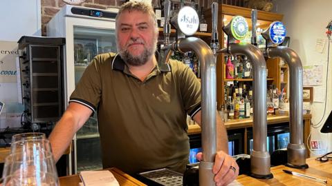 Daniel Bond, wearing a khaki polo shirt, poses behind the bar at his restaurant. He stands behind three beer pumps, next to the wooden bar top. 