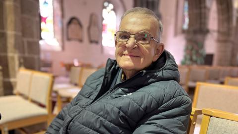 John smiles at the camera as he sits down on a chair in a church