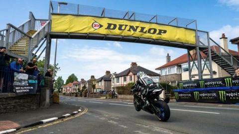 Michael Dunlop racing under the metal bridge with his front wheel off the ground. There is a large yellow Dunlop Tyres banner along the side of the structure overhead.
