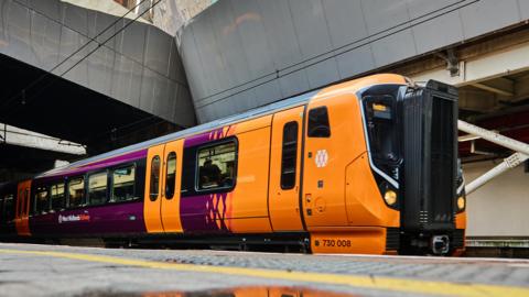 A West Midlands Railway train parked up at a railway station platform. It is orange and purple in colour with large windows and double doors. It has at least two carriages. On the platform is a puddle which reflects the train brightly