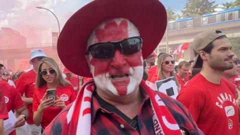 A man with his face painted to depict the Canadian flag leaf, wears a red hat and sunglesses.