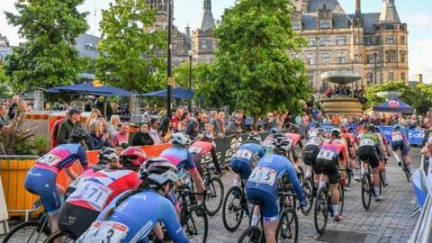 Many professional cyclists in lycra cycle down a temporary track which has been set up in a city centre. The town hall is visible in the background. Crowds of people line the route.