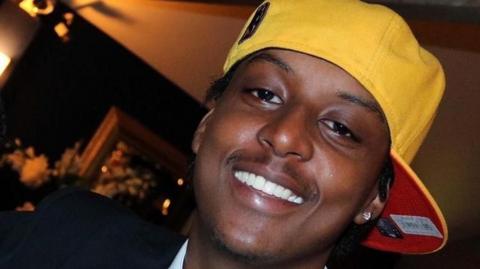 Wearing a yellow snapback hat, a close up photo shows Eddie Kinuthia smiling at the camera in a dark room.