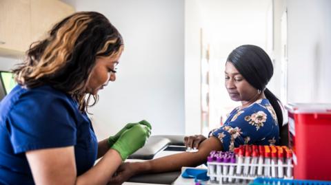 A  phlebotomists wearing green gloves taking the blood of a patient in a blue dress with flowers. They are sat in ward which has other blood samples and desk with a red box.