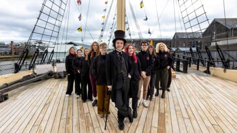An actor as Isambard Kingdom Brunel, wearing a top hat and black dress attire with a waistcoat, and a cain. He is standing in front of around 10 of volunteers wearing black SS Great Britain jumpers, all standing in a v formation. They are on the deck of the SS Great Britain, with the rigging and water in the background.