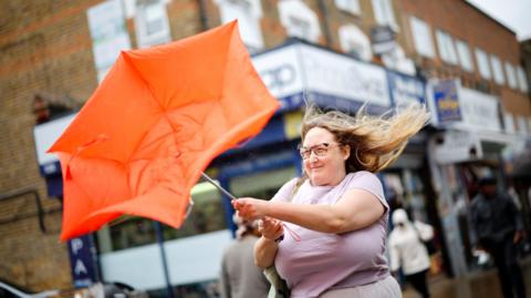 A windswept-looking woman holds an orange umbrella which has blown inside out with an urban street scene in the background.