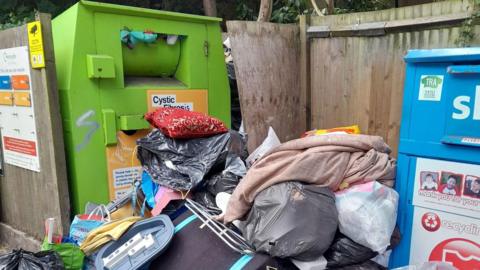 A pile of household waste, including black bags, full carrier bags and other items. The pile is between a large green recycling bin and a similar blue bin, and surrounded by wooden fencing.