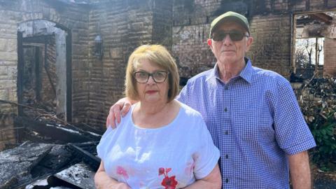 A woman with brown hair, glasses and a white patterned T-shirt next to a man with a green cap, blue shirt and sunglasses with one arm over her shoulder and the remains of their fire-damaged home behind them