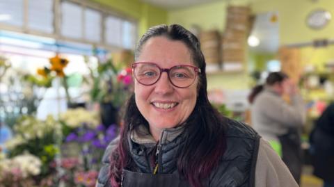 Michelle Bly who is wearing an apron with rows of flowers behind her in her shop. She has brown hair and glasses and smiling at the camera