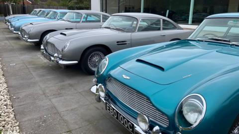 Six Aston Martin DB5s can be seen parked in a line in front of a glass fronted building. The nearest car to to the camera is a green-blue colour. They are parked on grey paving slabs.