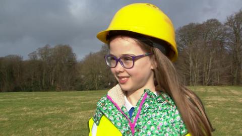 A young girl with long brown hair is smiling at someone off to the side of the camera. She is wearing blue framed glasses and a yellow hard hat. Her coat is green patterned with a pink zip. She is standing in a grassy area with trees in the background.