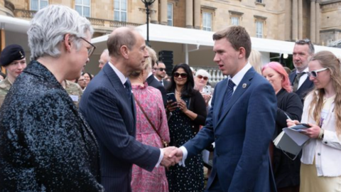 Prince Edward can be seen talking and shaking hands with Jacob. They are in formal wear and surrounded by a crowd of people at Buckingham Palace.