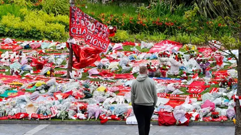 A man, with his hands in his pockets, looks at flowers, flags and football shirts laid outside Anfield for Diogo Jota