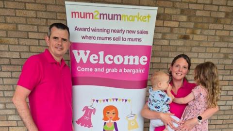 Emmie Withers is smiling at the camera, wearing a pink t-shirt and holding her two children is standing by a mum2mum market sign with her husband also wearing a pink shirt.
