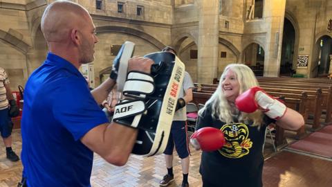 Boxing fitness coach Ben Cookson holds up pads while a woman with red and white boxing gloves and a Cobra Kai T-shirt throws a punch. The class is inside a church.