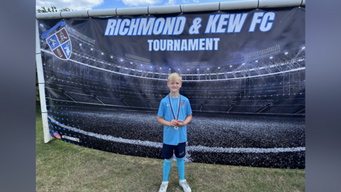 A boy with blond hair, wearing a football blue jersey and black shorts, long socks and white trainers. He is standing in front of a large black banner. He is wearing a medal around his neck.