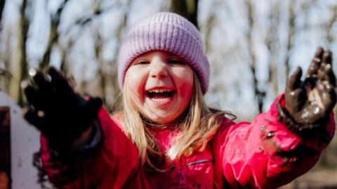 A young girl with blonde hair wearing a lilac woolly hat and pink waterproof coat holds out her hands which are full of mud. She is laughing. She is standing in WWT Martin Mere Wetland Centre with trees in the background.