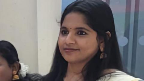 Renjitha Gopakumaran Nair stands in a room with another woman beside her. She is smiling, has long, straight dark hair and is in traditional dress.
