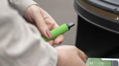 File image showing a woman holding a green disposable vape in her hand while she throws the cardboard packaging for it in a black street bin in London