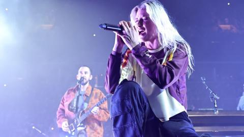 Mike Shinoda and Emily Armstrong of Linkin Park perform in New York. Emily, in the foreground, closes her eyes and kneels as she sings into a mic, held in both hands. She has bleached blonde hair worn loose and wears a white and purple sweatshirt. Mike, in the background, plays guitar and sings while wearing an orange jacket. 