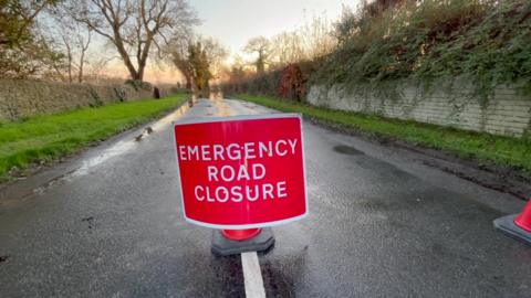 Emergency Road Closure sign on a road with grass verges either side.