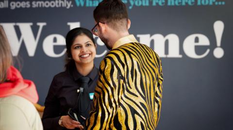 A woman and man in conversation in front of a sign that reads 'Welcome!'