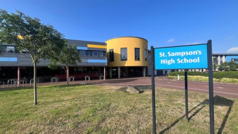 St Sampson's High School. A sign at the front of the building with a yellow, blue and grey building behind