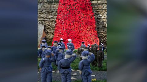 A waterfall of red poppies on a stone wall with small knitted soldiers on the grass in front of it 