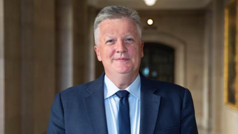A man with short grey hair in a navy suit with a blue shirt and tie