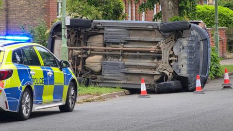 A police car parked on a road in front of a car that has overturned and is surrounded by orange traffic cones. 