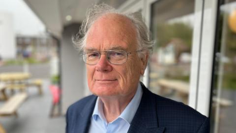 A man called Peter Harwood. He is elderly and has silver-rimmed glasses and whispy grey hair. He is wearing a navy blaze with a blue shirt underneath.