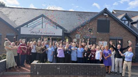 A crowd of staff stand outside the single-storey Teesside Hospice building and let off confetti streamers while cheering. Lots of staff have their hands held in the air and they are all smiling. Jets of confetti are floating in the air.