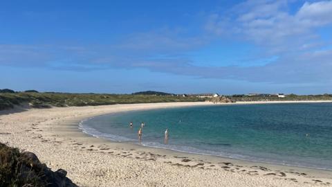 Port Soif Bay in Guernsey. It is a large sweeping bay with white sands and bright blue sea. Grassy headland stretches around the top of the sand. People are swimming and walking into the sea. 